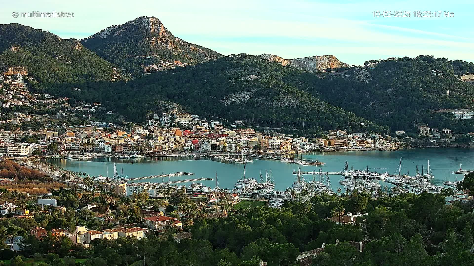 A coastal town nestled between verdant hills and a calm bay filled with numerous sailboats, under a clear sky.