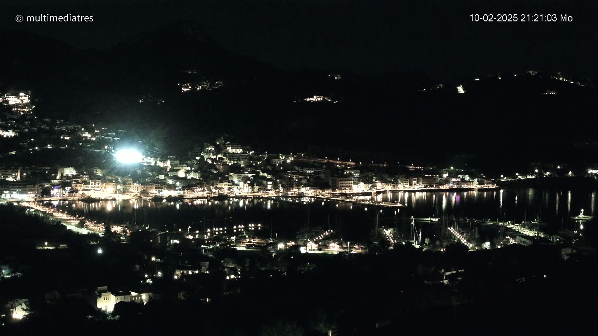 A nighttime, aerial view shows a harbor town brightly lit along a calm body of water, nestled between dark hills.
