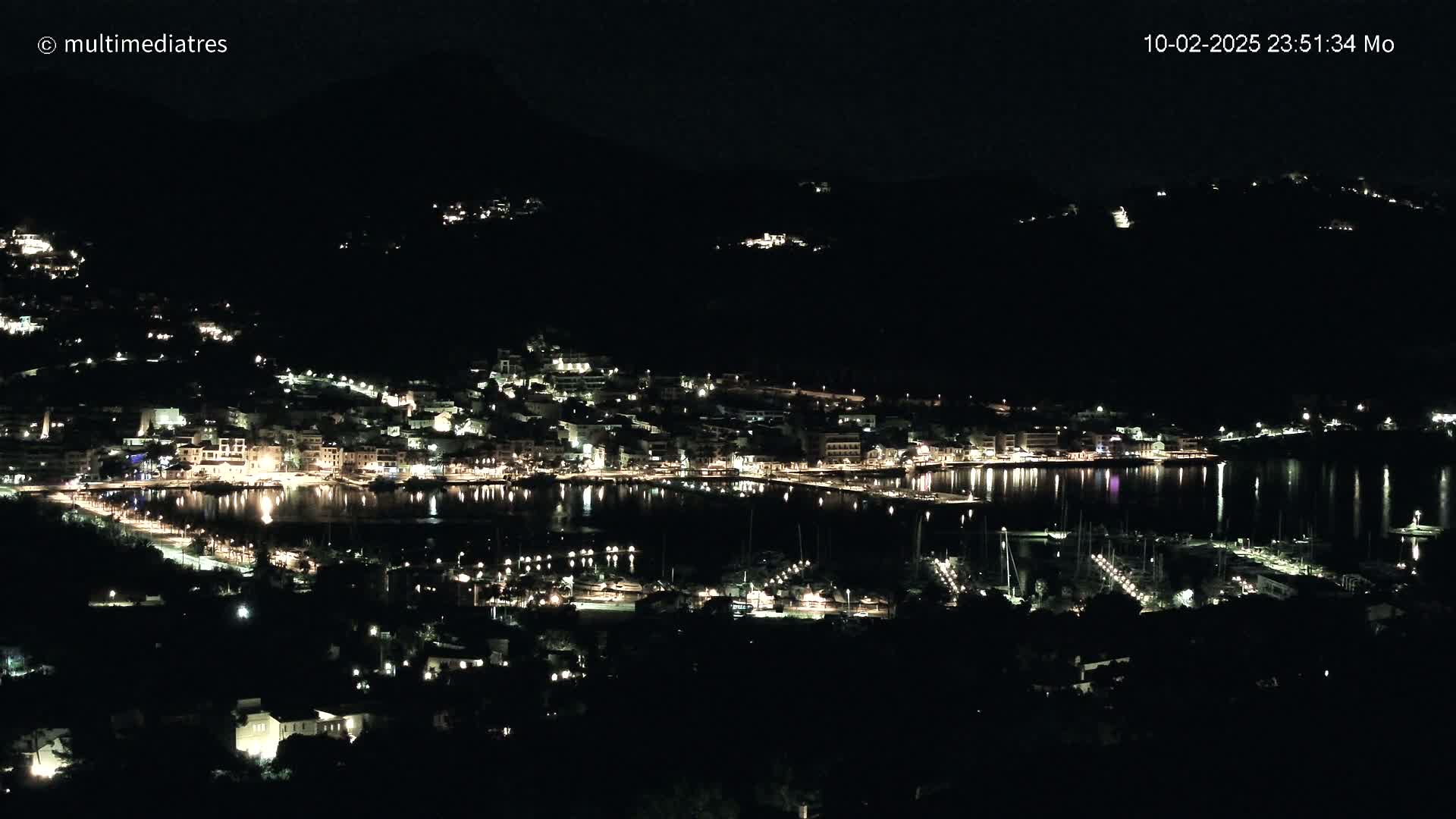 A nighttime view of a coastal town, illuminated with lights, and a calm body of water reflecting the lights.