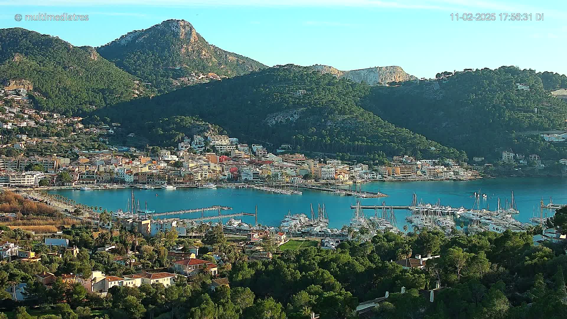 A coastal town nestled between green hills and a calm, blue bay filled with numerous sailboats under a clear sky.