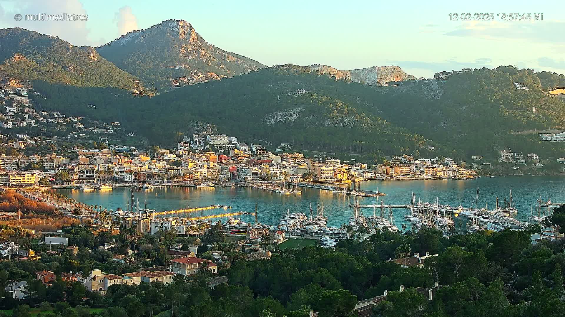 A coastal town nestled between mountains with a calm harbor filled with sailboats under a clear evening sky.
