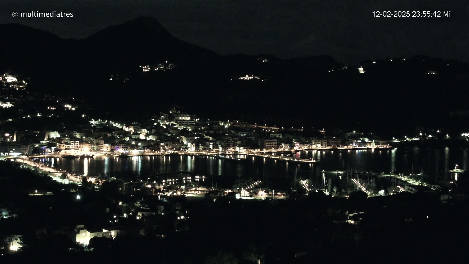A nighttime view of a coastal town illuminated by lights, nestled between dark hills and a calm, reflective body of water filled with boats.