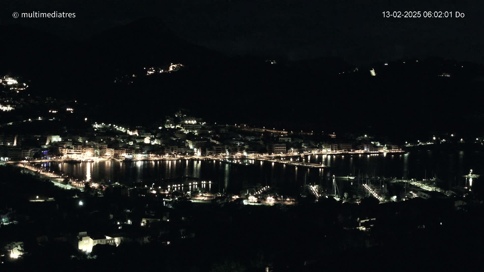 A nighttime view of a harbor town nestled against a dark hillside, illuminated by the lights of buildings and boats.