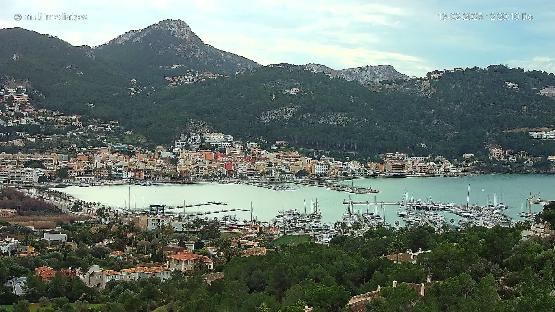 A coastal town nestled at the base of a mountain range on a partly cloudy day, featuring a marina filled with numerous boats.
