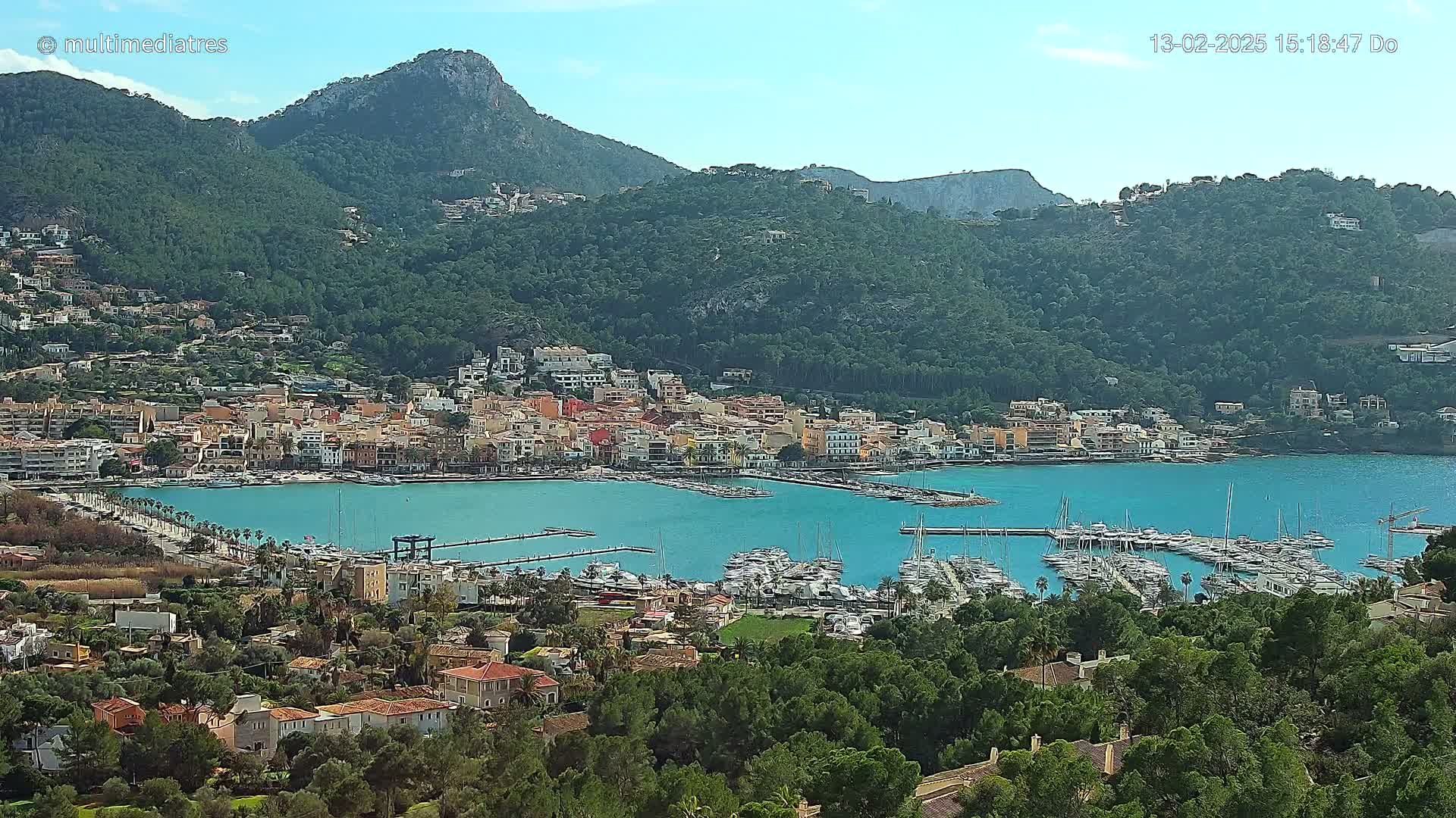 A coastal town with a marina full of boats sits nestled at the base of a mountain range on a clear, sunny day.