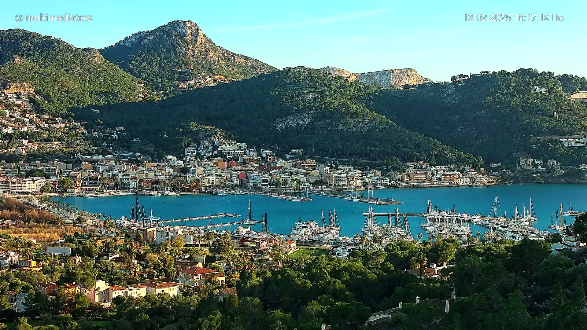 A coastal town nestled between mountains, featuring a harbor filled with sailboats, under a clear blue sky.