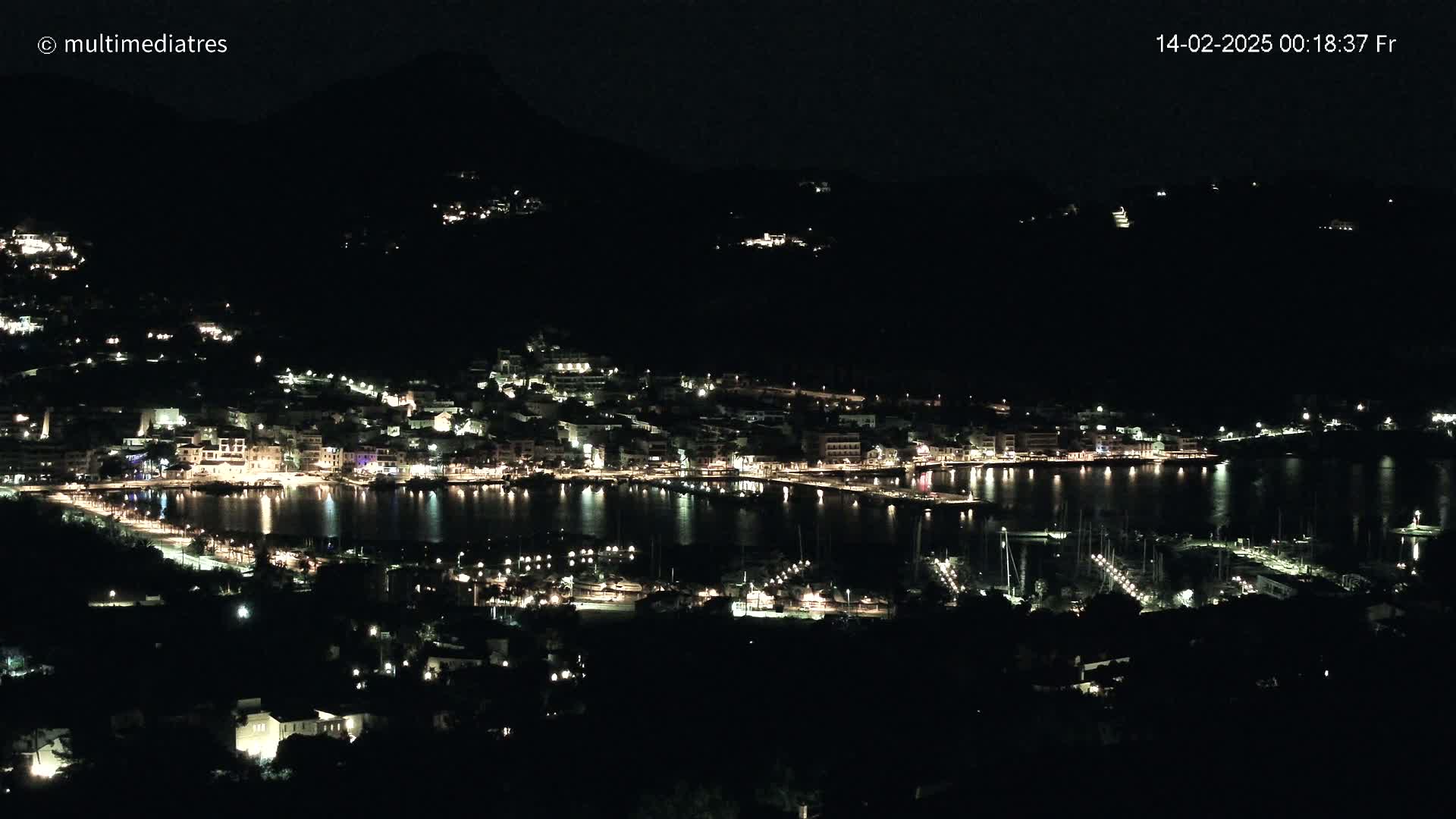 A nighttime, aerial view of a coastal town illuminated by lights, nestled against a dark hillside and featuring a harbor filled with boats.