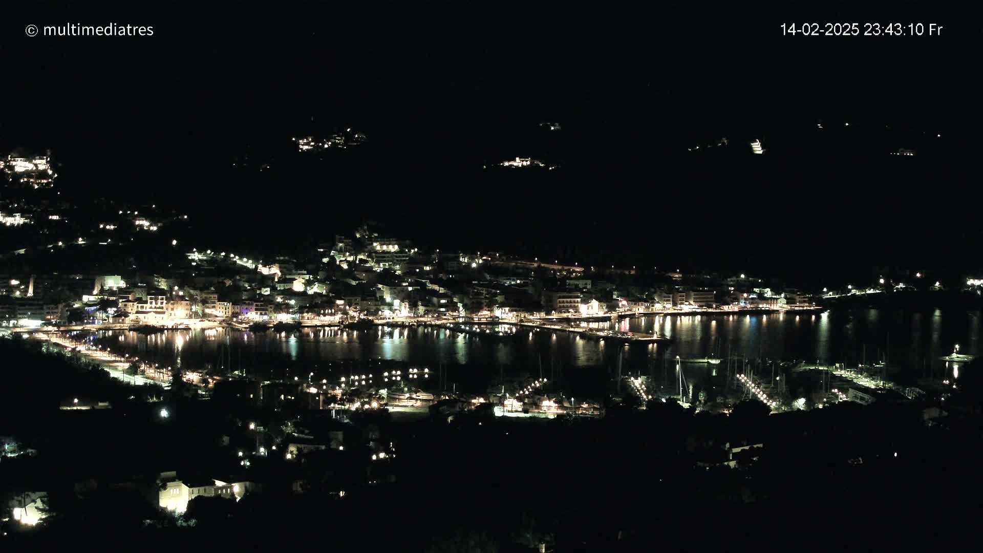 A nighttime view of a harbor town, with its buildings and boats illuminated, situated on a dark hillside under a clear night sky.
