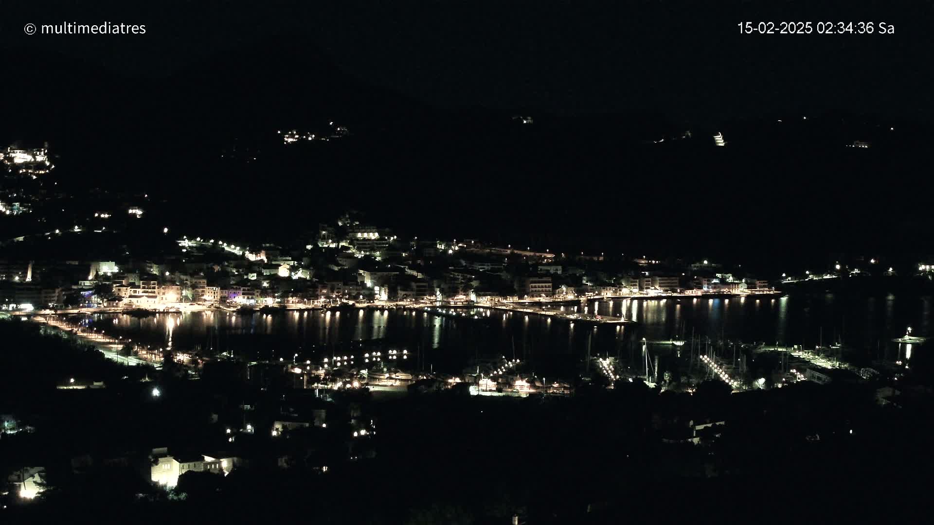 A nighttime view of a coastal town with its lights reflected on calm water, surrounded by dark hills.