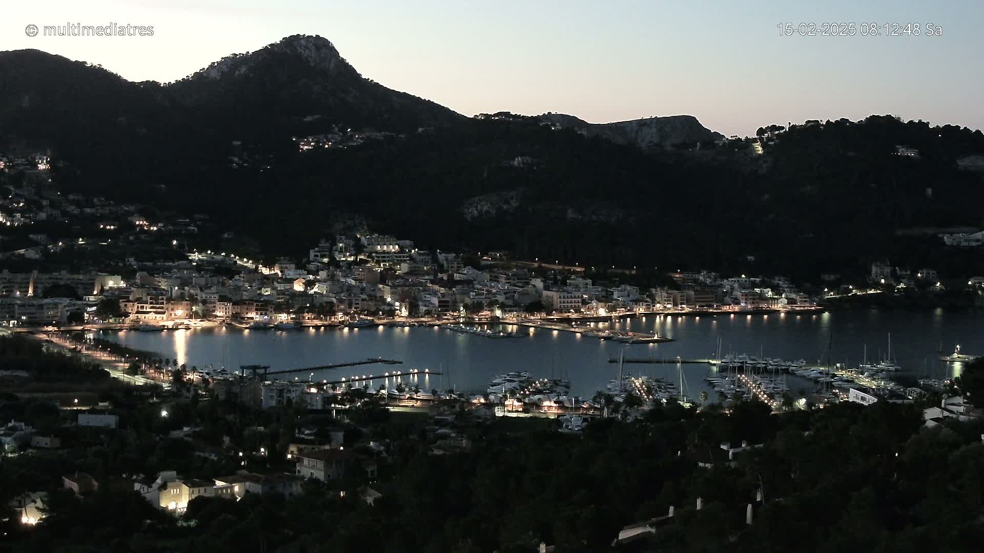 A nighttime view of a harbor town nestled below a dark mountain, illuminated by lights, under a twilight sky.