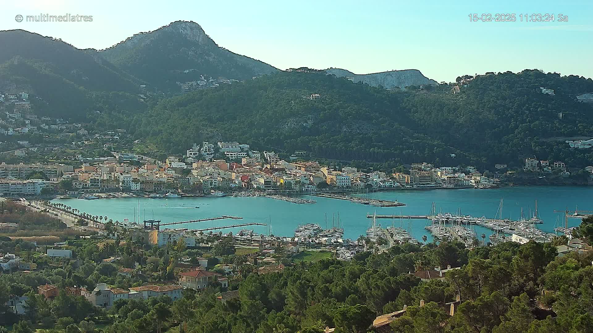 A coastal town nestled between green hills and a calm bay filled with numerous boats under a clear sky.