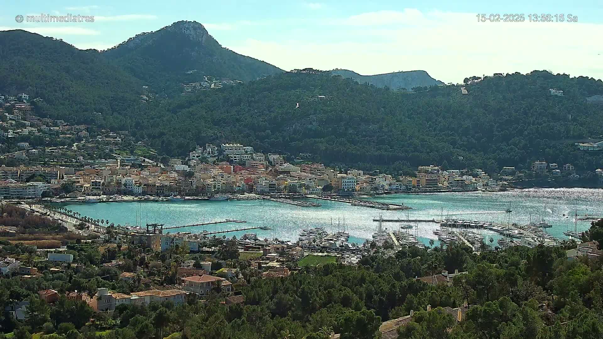 A coastal town nestled in a mountainous area with a harbor filled with boats under a mostly sunny sky.