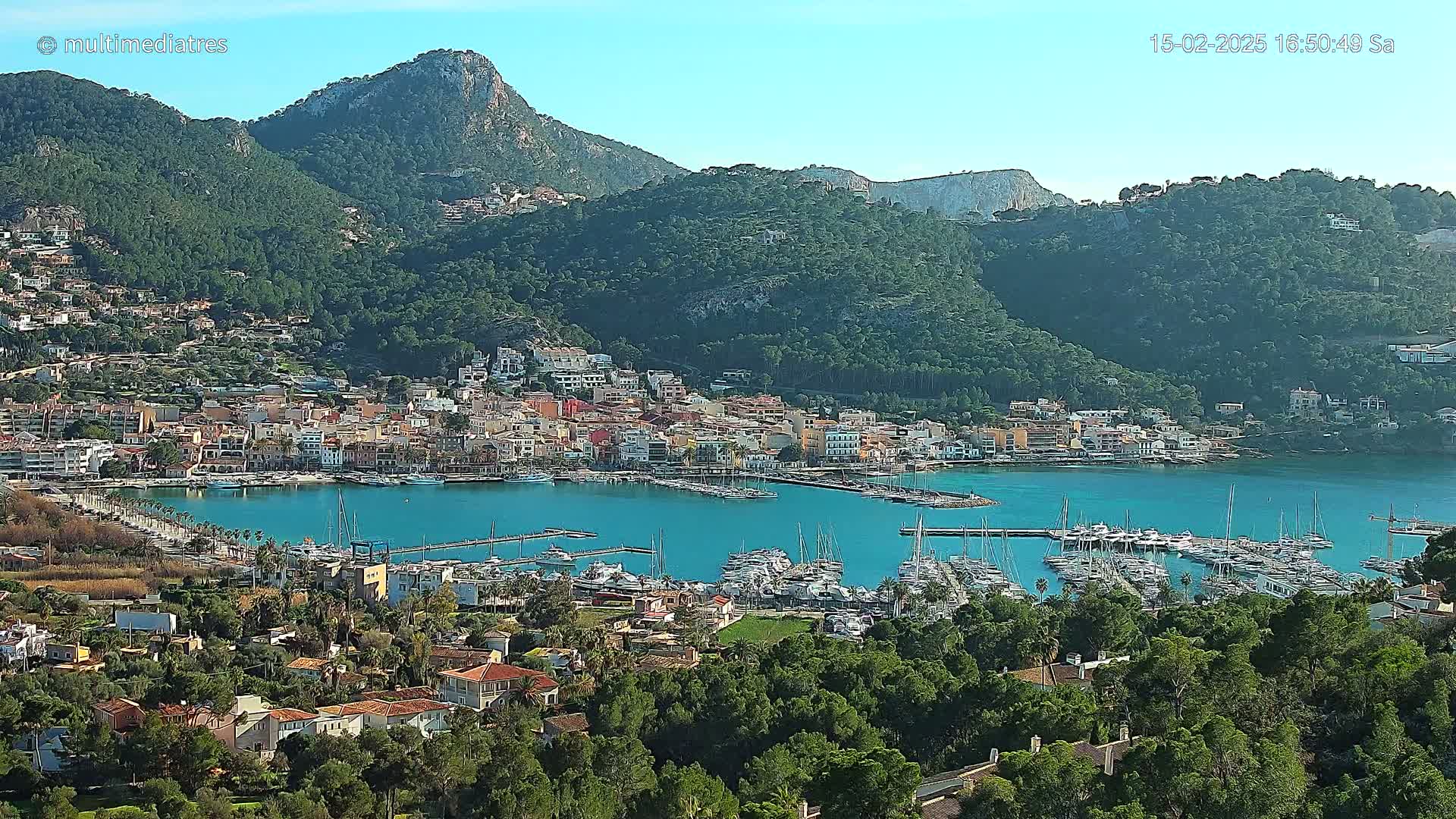 A picturesque coastal town nestled between verdant hills and a calm, turquoise harbor filled with numerous boats under a clear, sunny sky.
