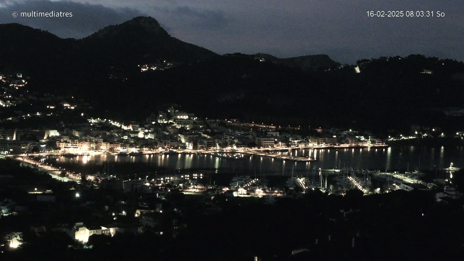 A nighttime view of a coastal town nestled between dark hills, illuminated by many lights, with a calm harbor filled with boats.