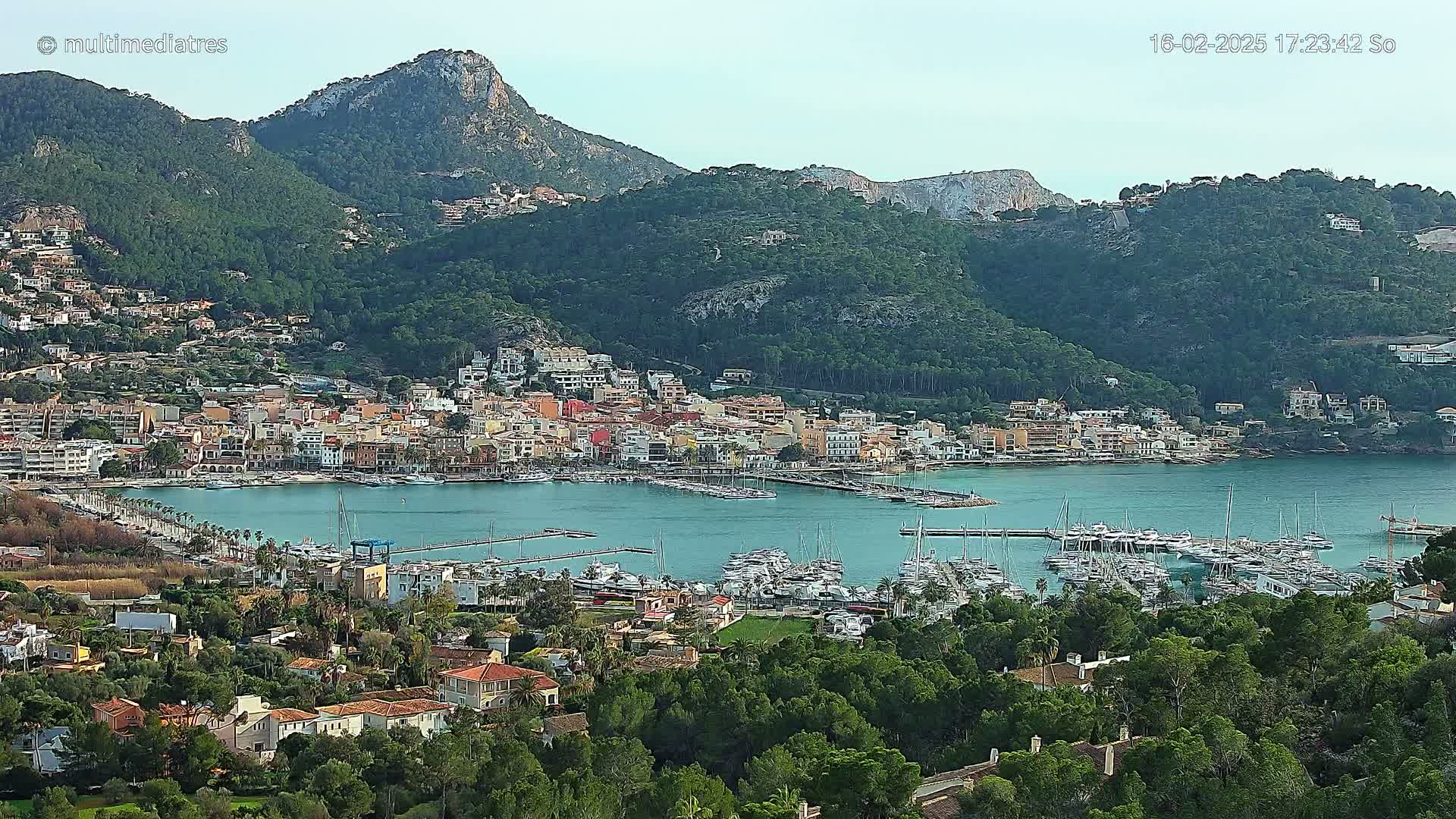 A coastal town nestled between green hills and a calm bay filled with numerous boats on a clear day.