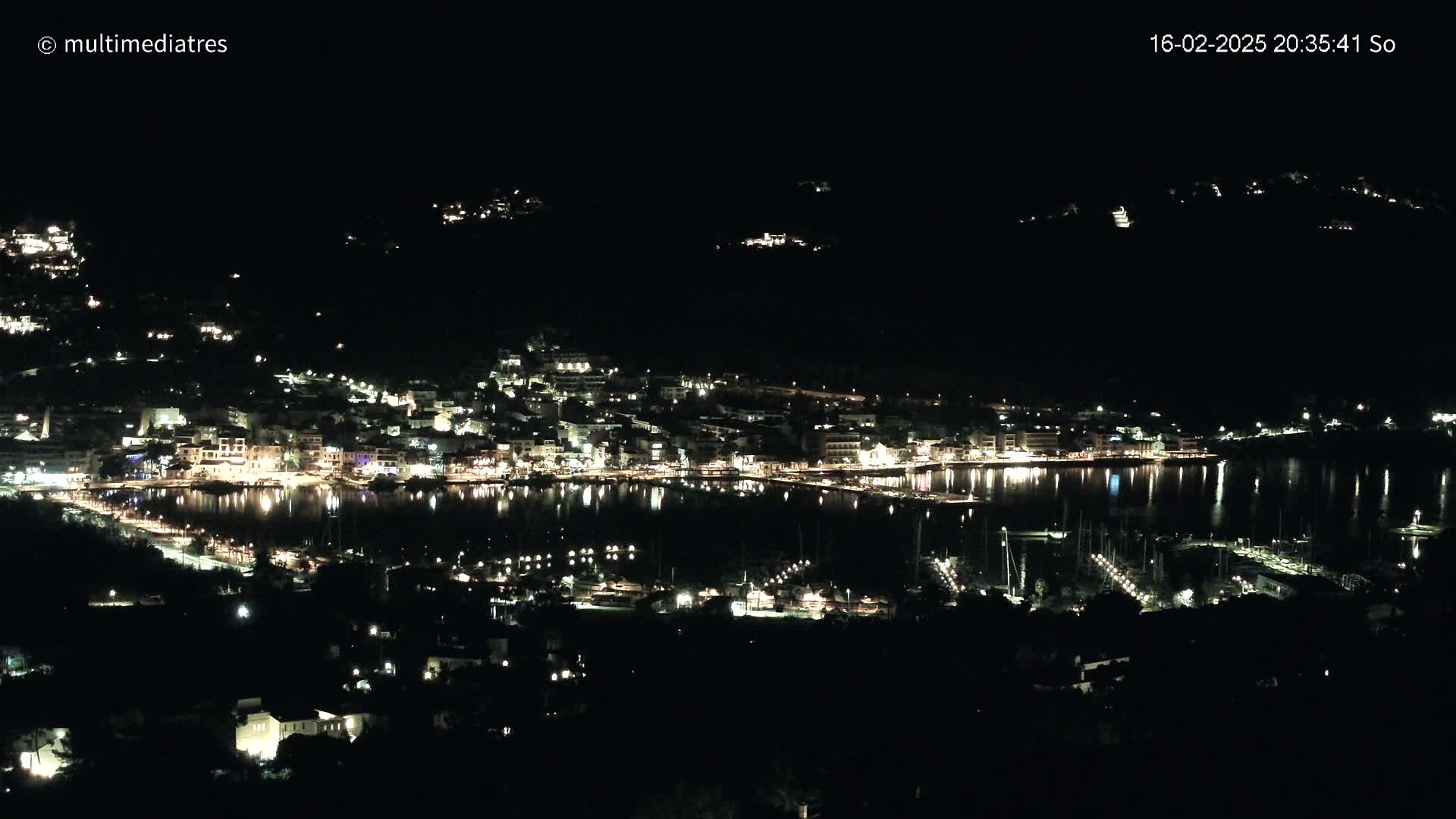 A nighttime, aerial view shows a coastal town with its lights reflecting on a calm body of water, where many boats are docked.