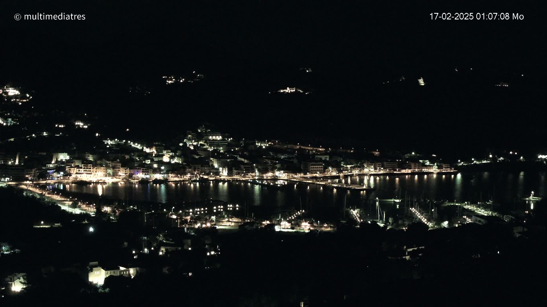 A coastal town is brightly lit at night, its lights reflected in the calm water of a harbor filled with boats.
