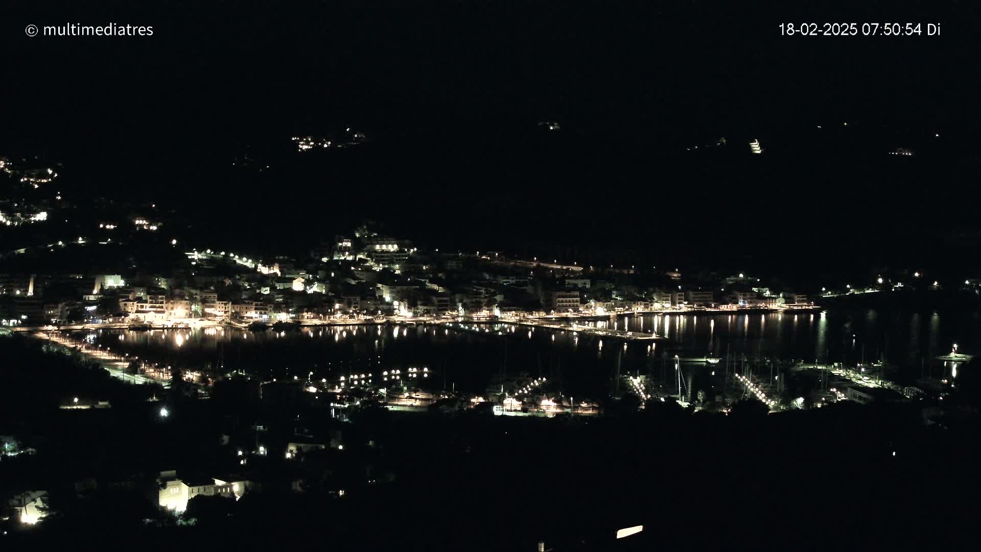 A nighttime view of a coastal town illuminated by lights, with a calm body of water reflecting the lights.