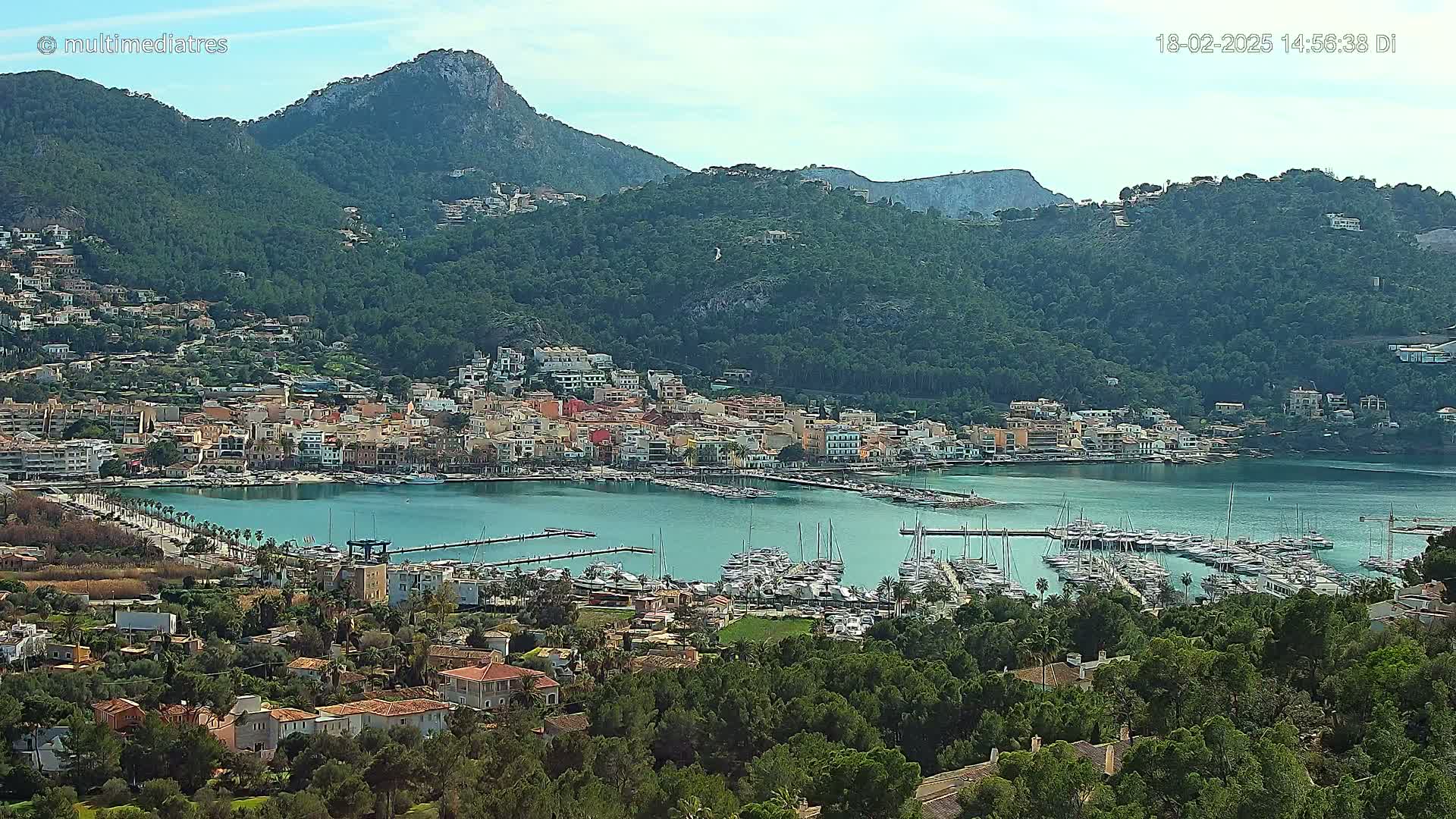A coastal town with a marina filled with boats sits nestled at the base of a verdant mountain range under a clear sky.