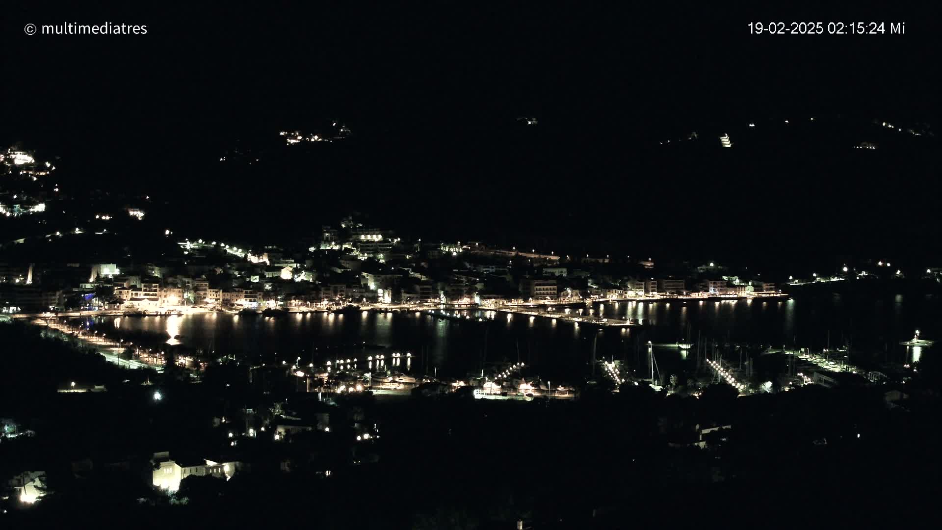 A coastal town is illuminated at night, with its lights reflecting on a calm body of water, under a clear, dark sky.