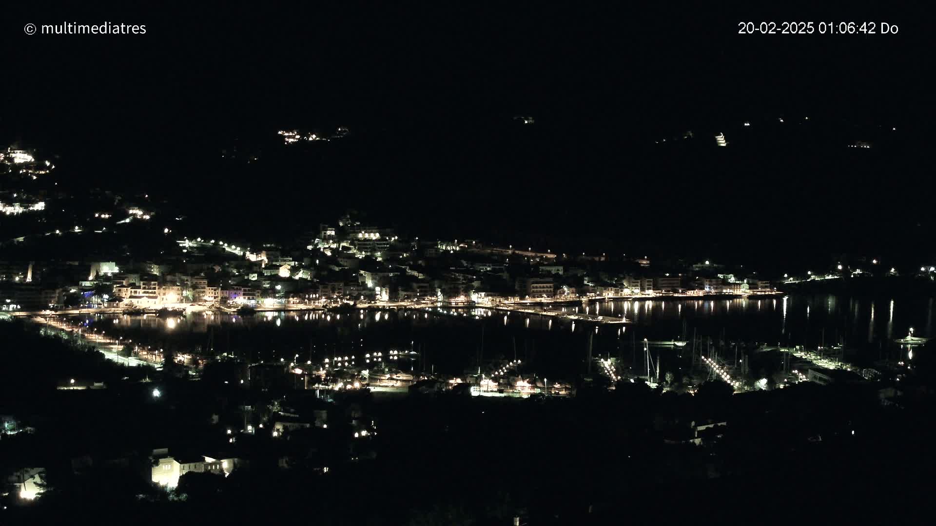 A coastal town at night is illuminated by many lights along its waterfront, reflecting on the calm water of the harbor.