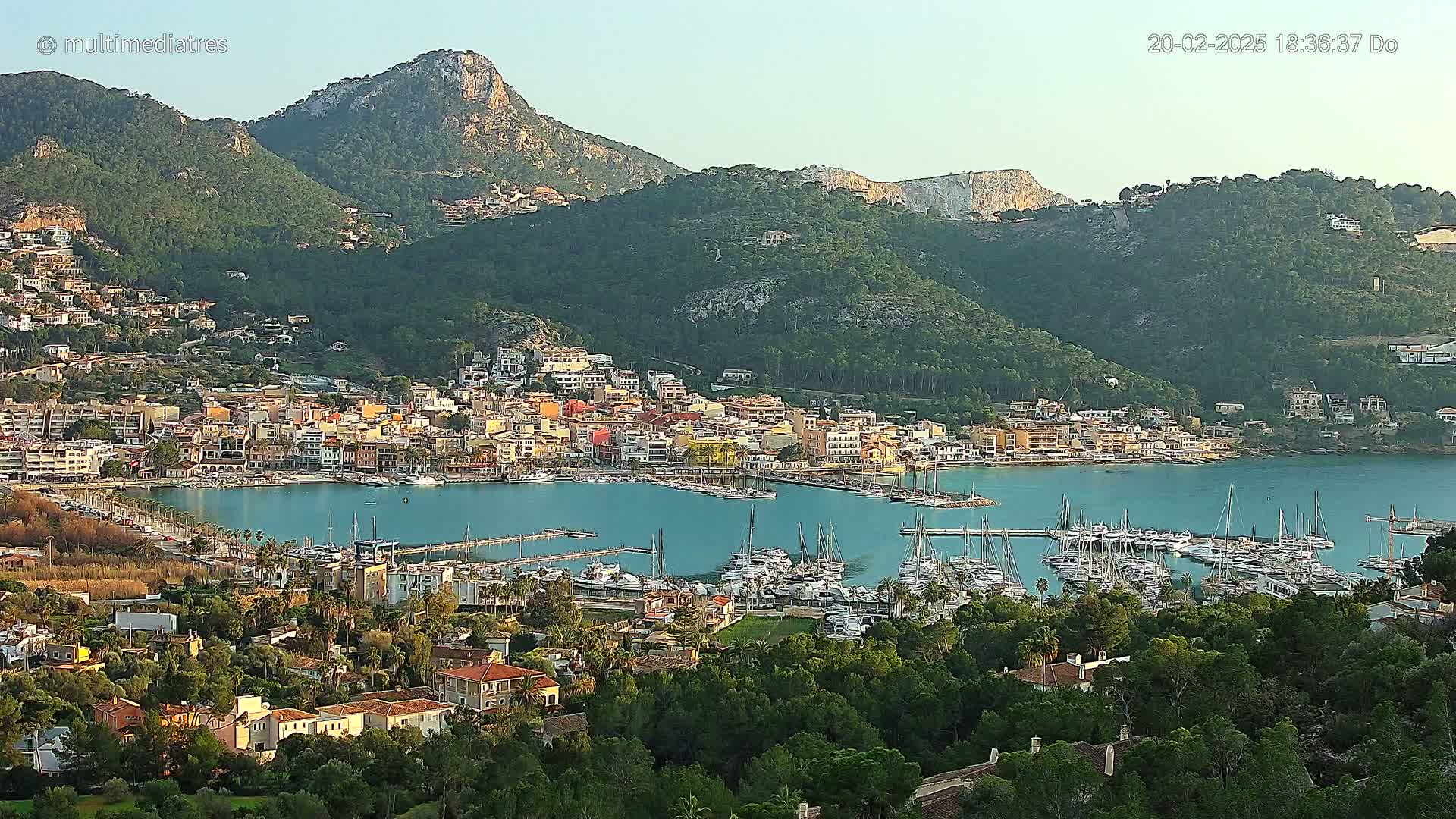 A coastal town nestled in a mountainous region, with a marina filled with sailboats and clear, calm water under a mostly clear sky.