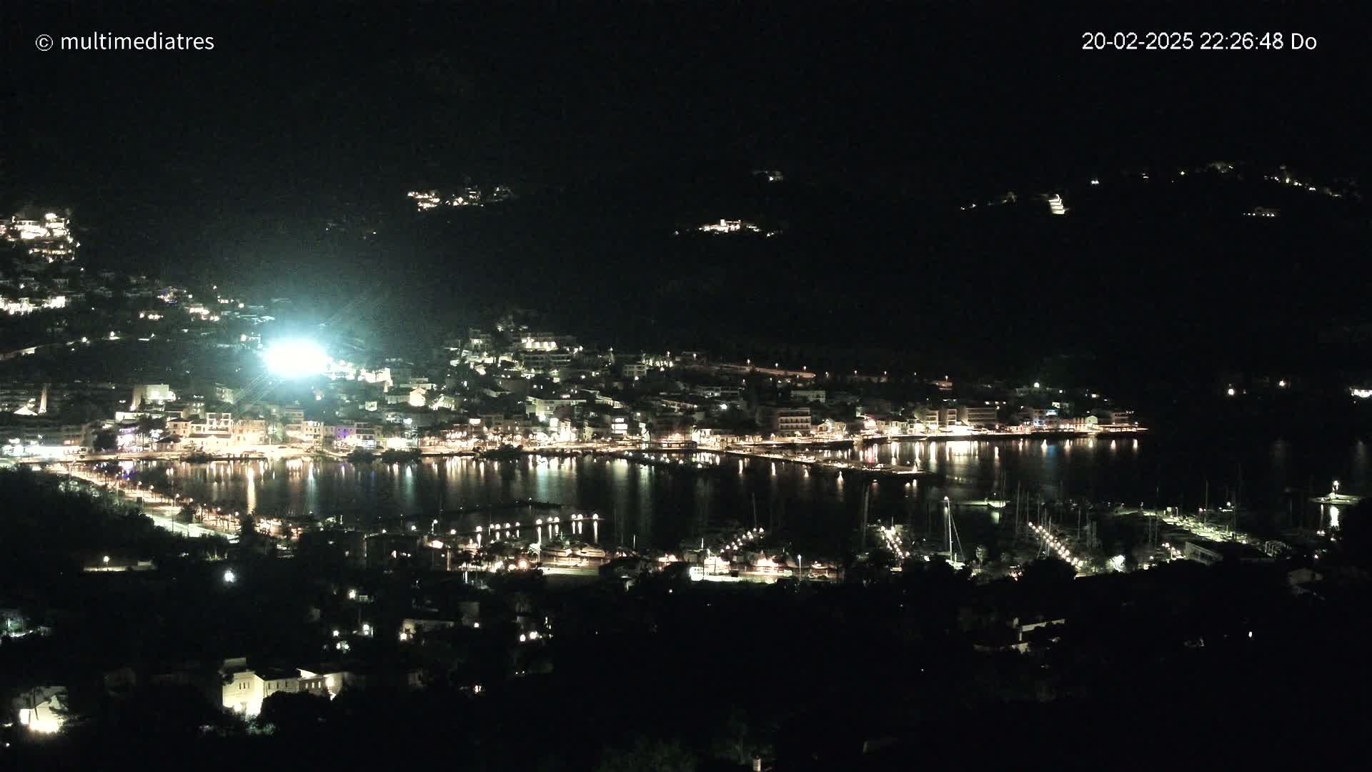 A nighttime view of a harbor town, illuminated with lights, nestled along a dark, calm body of water under a clear, dark sky.