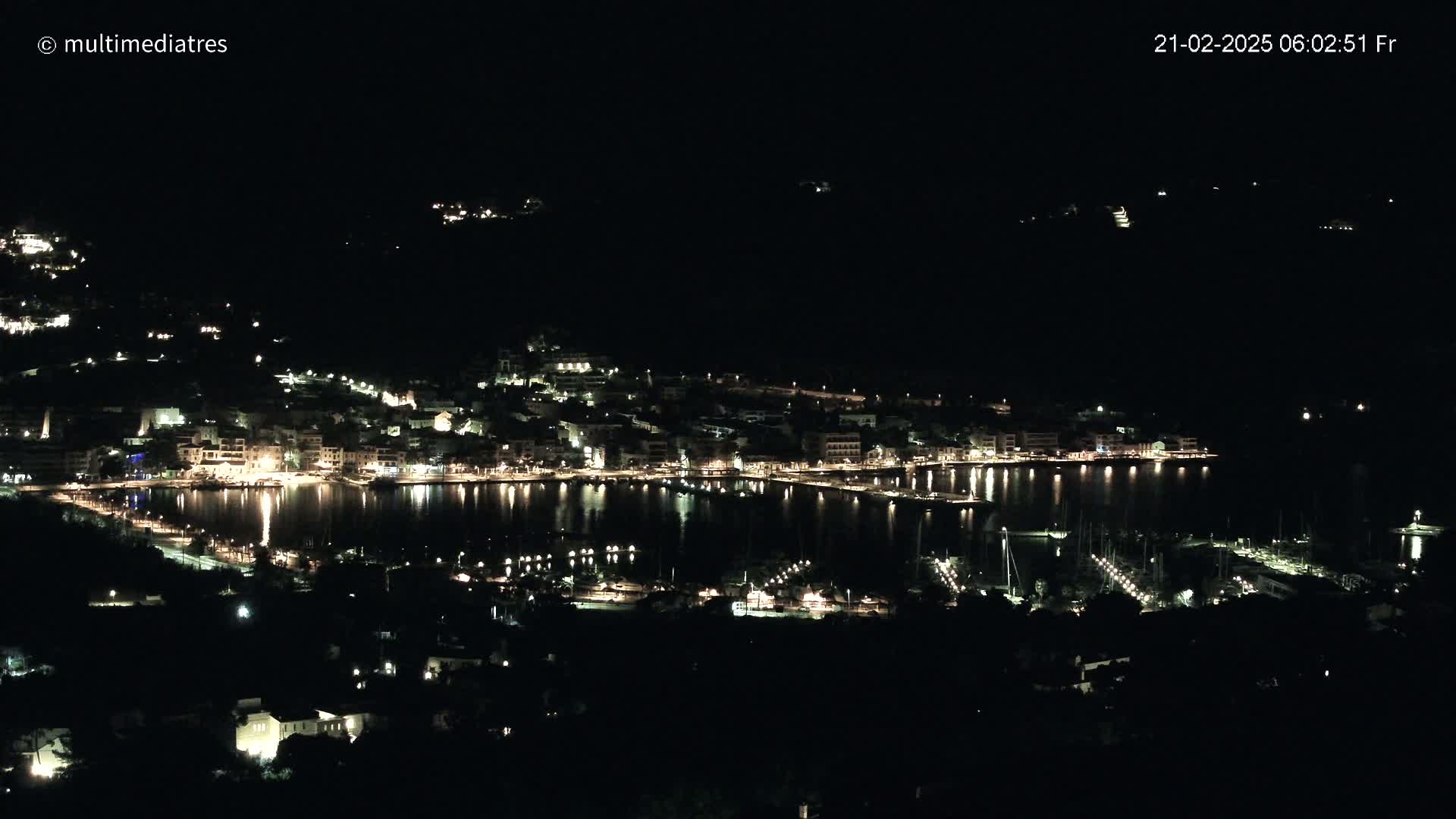 A nighttime, high-angle view shows a coastal town with its lights reflecting on a calm harbor, surrounded by dark hills.