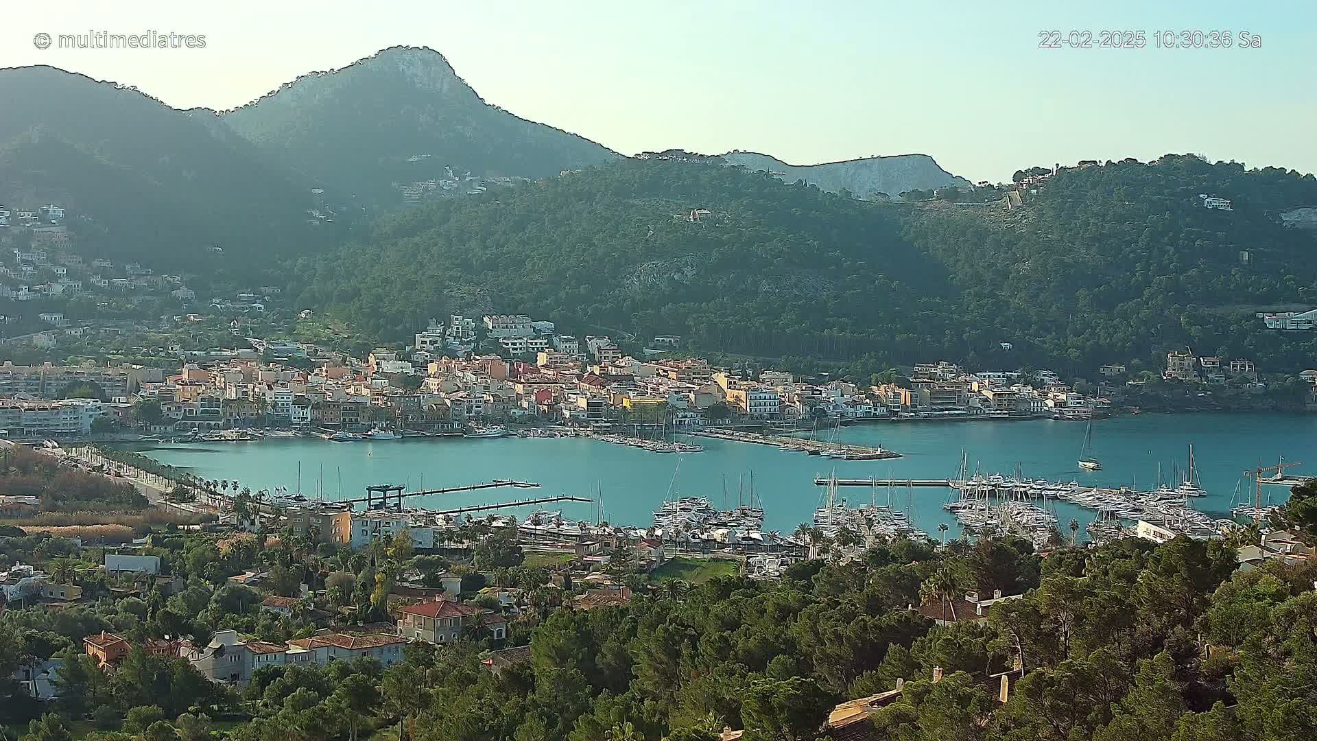 A coastal town nestled between verdant hills and a calm bay filled with numerous sailboats under a clear sky.