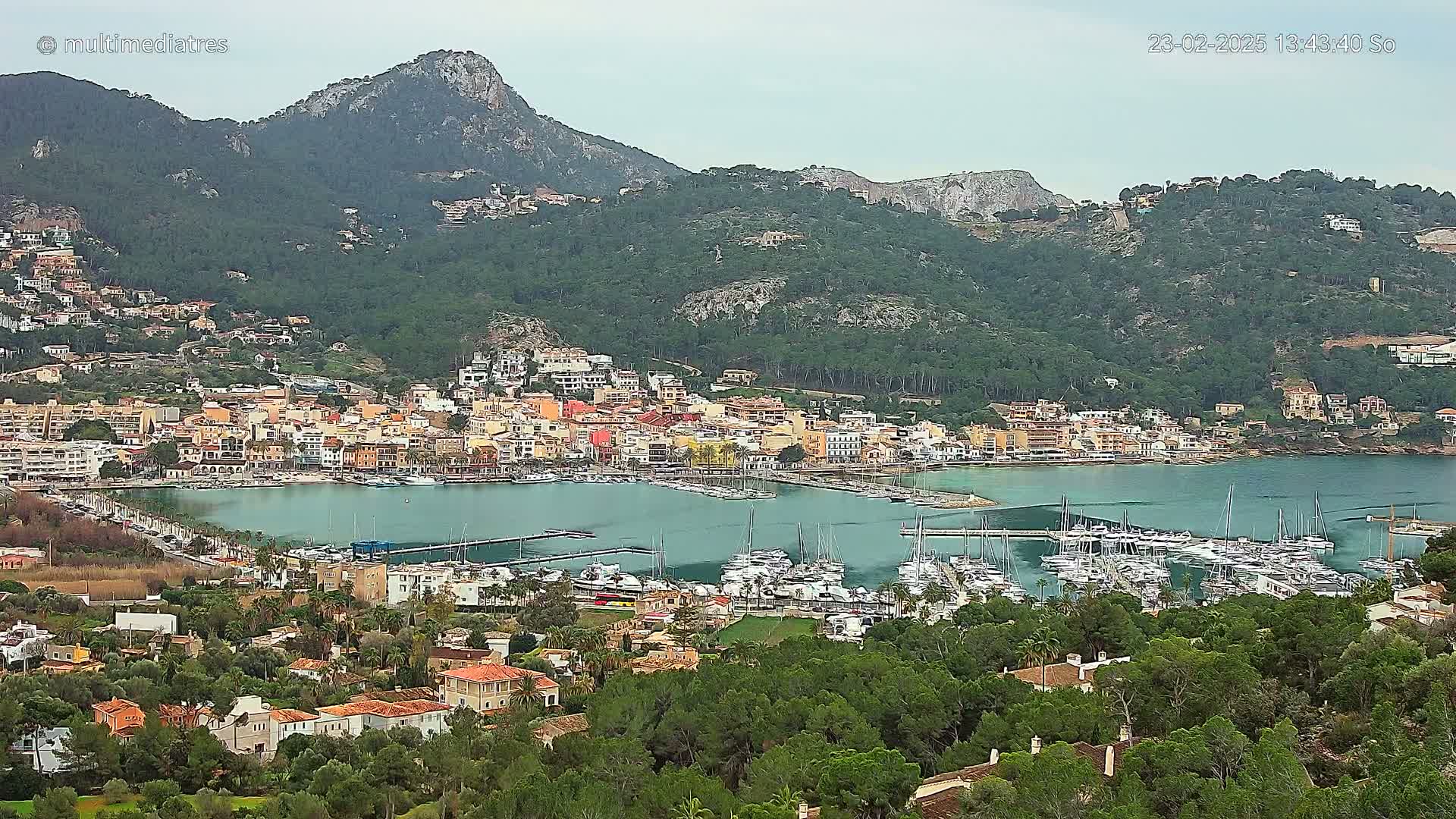 A picturesque coastal town nestled at the base of a mountain range, featuring a marina filled with sailboats under an overcast sky.