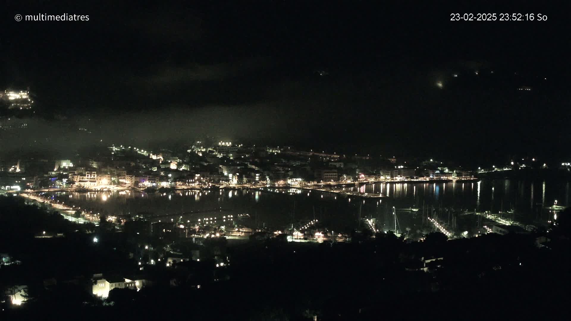 A nighttime, aerial view of a coastal town with a harbor filled with boats, under a slightly hazy sky.