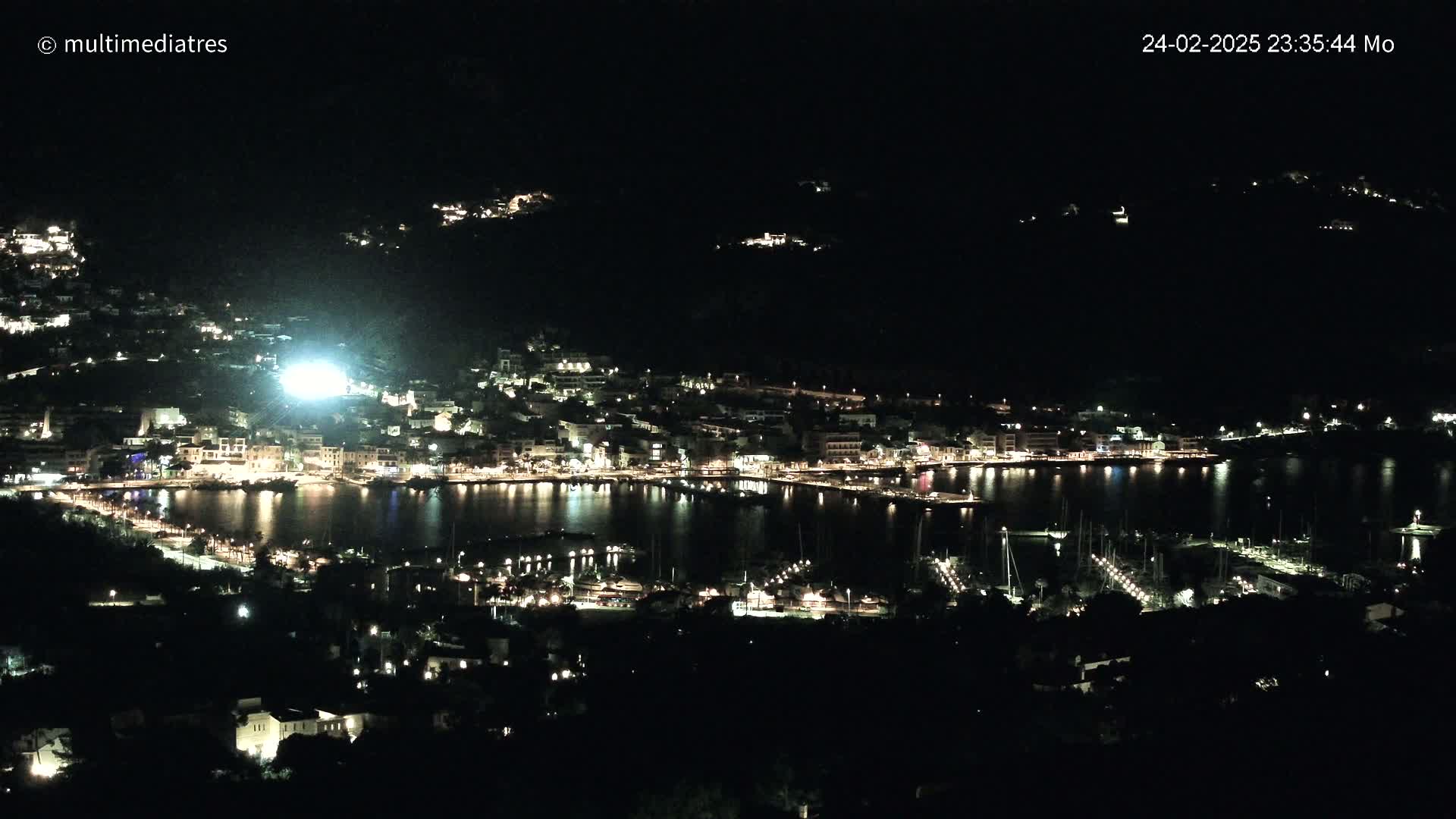 A nighttime, aerial view of a coastal town with a harbor full of boats, illuminated by many lights.