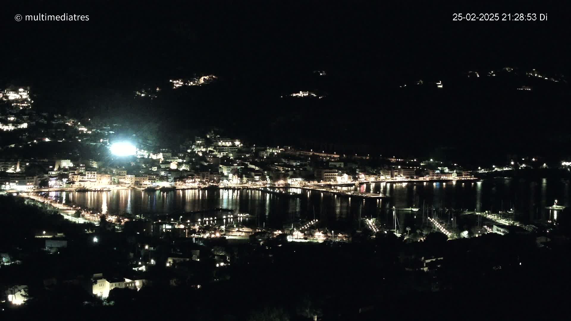 A nighttime view of a harbor town illuminated by lights, nestled against dark hills under a clear, dark sky.