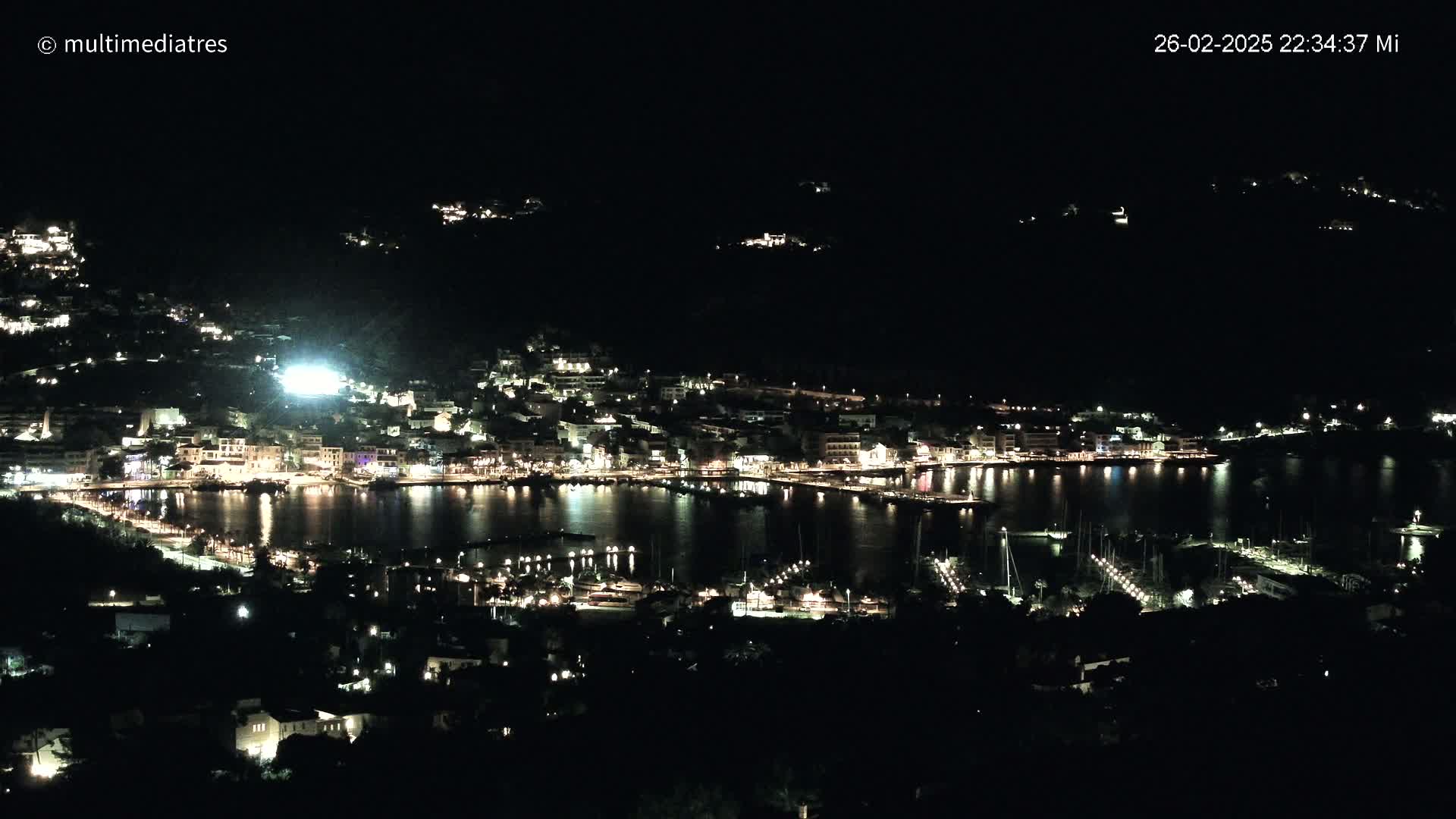 A nighttime view of a coastal town with its lights illuminating the harbor and surrounding hills.