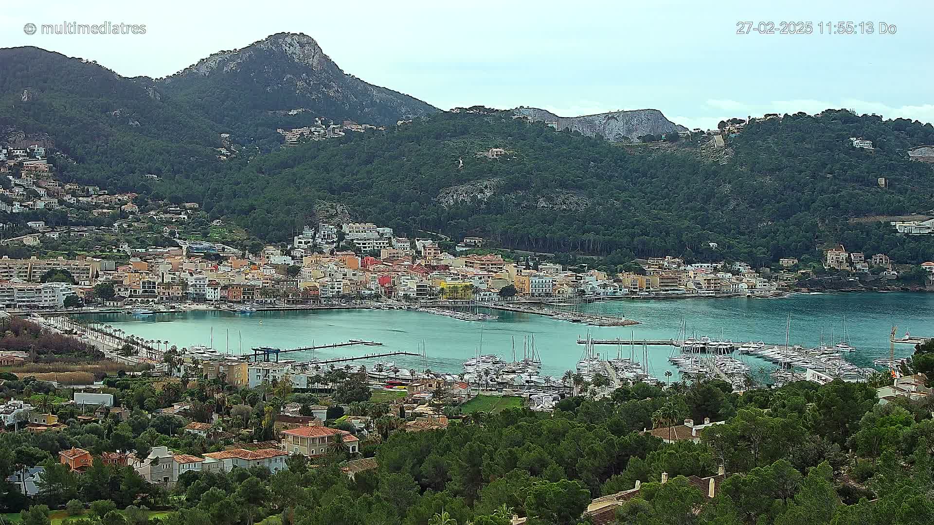 A coastal town with a marina full of sailboats nestled at the base of a mountain range under an overcast sky.