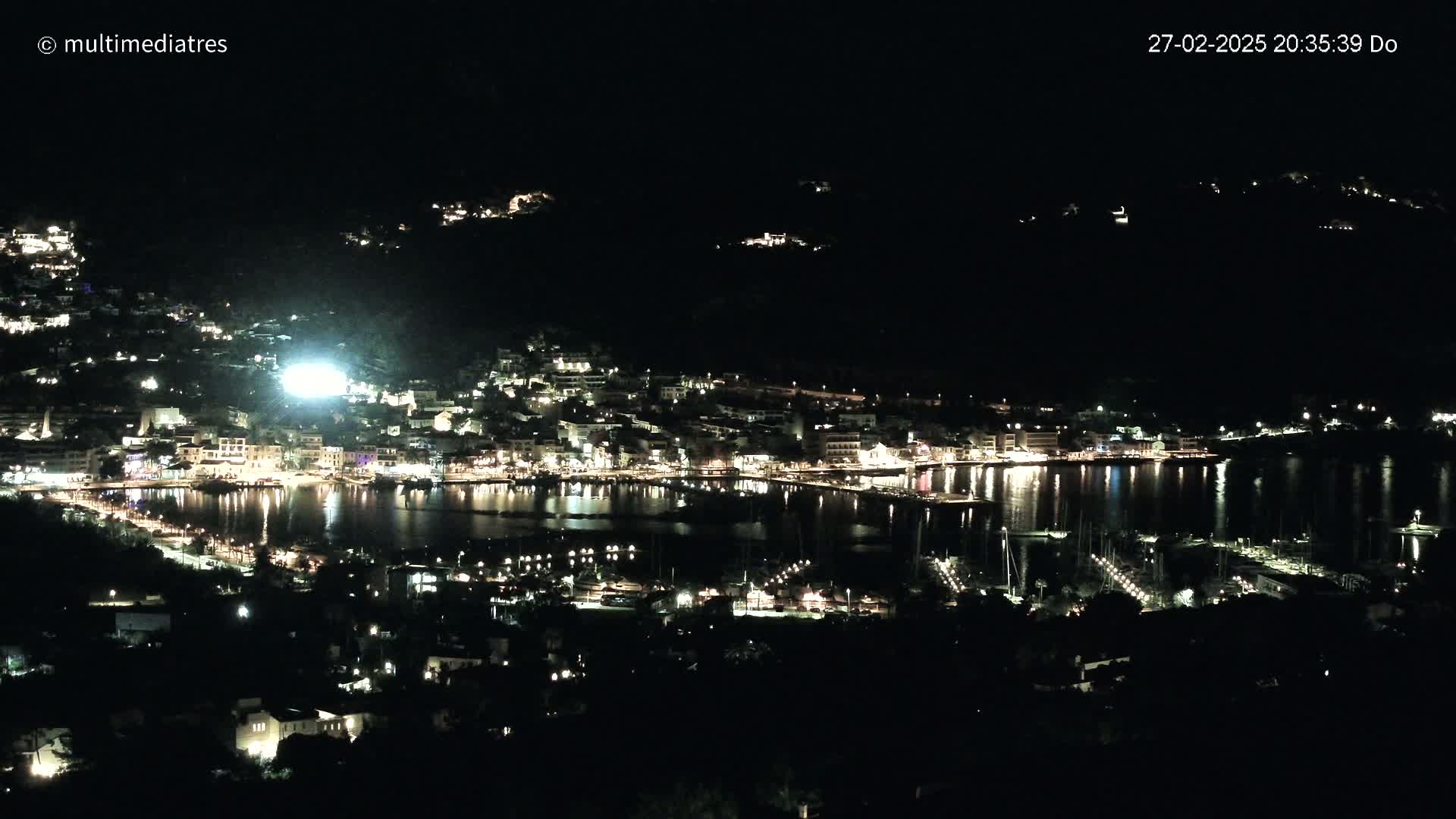 A nighttime, high-angle view of a coastal town illuminated by many lights, with a calm body of water reflecting the lights.