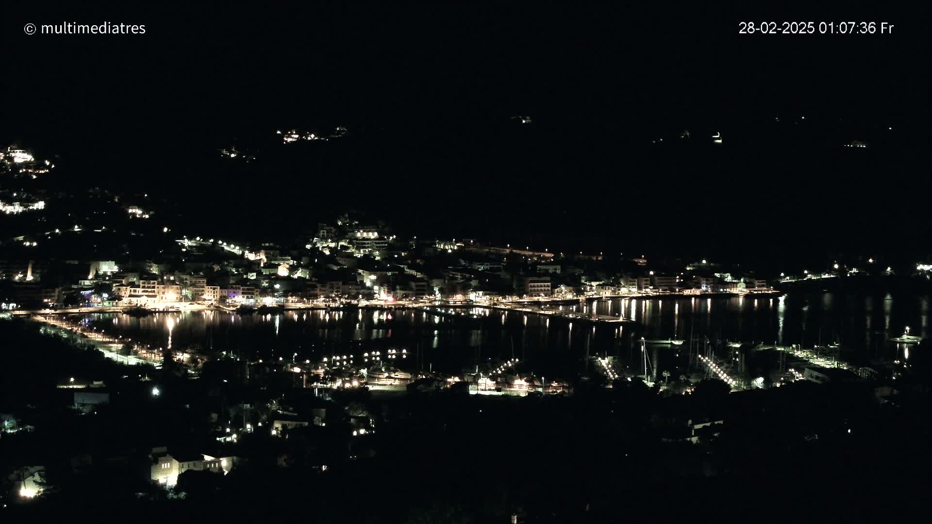 A coastal town at night, illuminated by numerous lights reflecting on the calm water of a harbor.