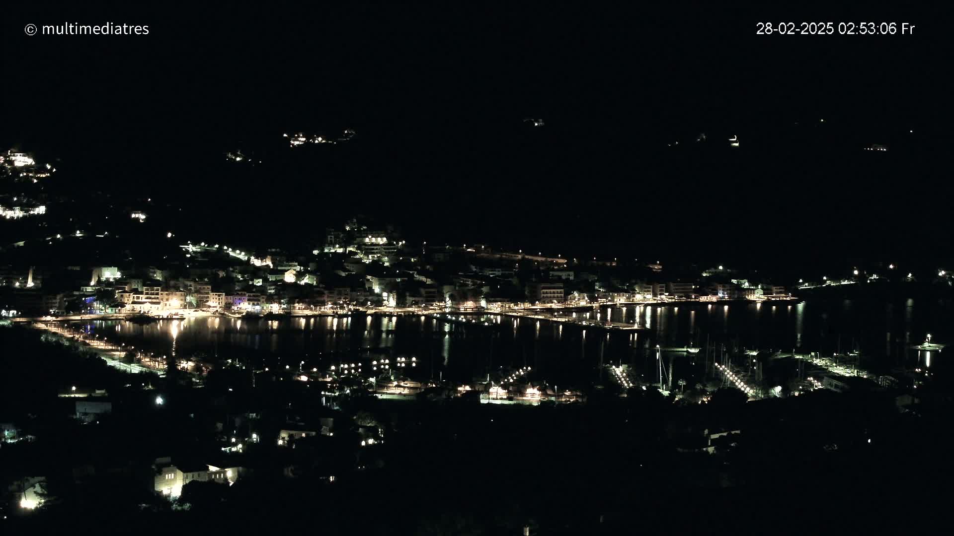 A nighttime, aerial view of a coastal town with its lights illuminating the waterfront and surrounding buildings.