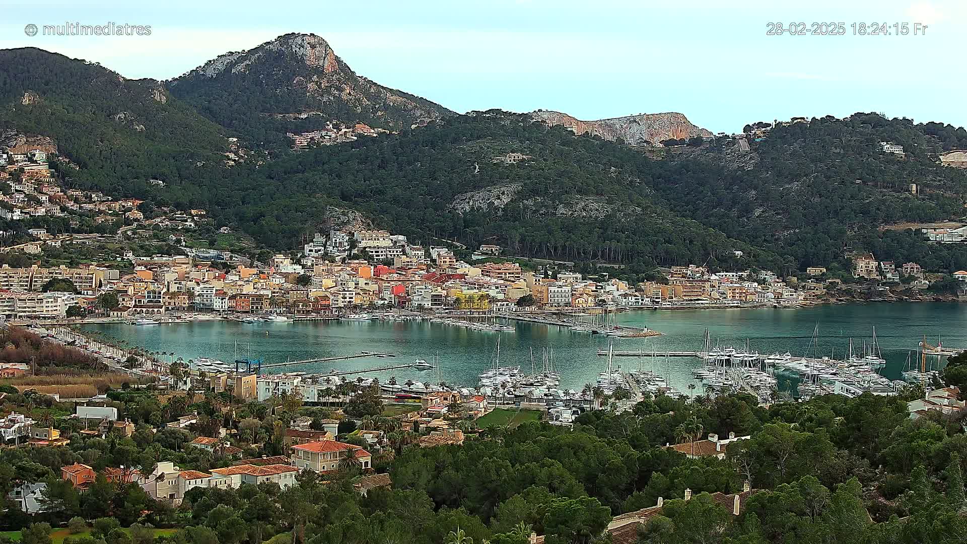 A harbor filled with sailboats is nestled in a town of colorful buildings at the base of a verdant mountain range under a mostly clear sky.