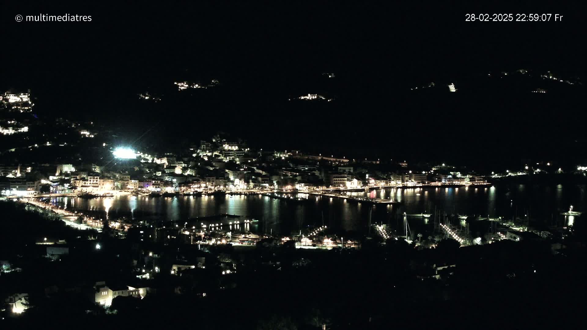 A nighttime view of a harbor town illuminated with lights, nestled against dark hills under a clear, dark sky.