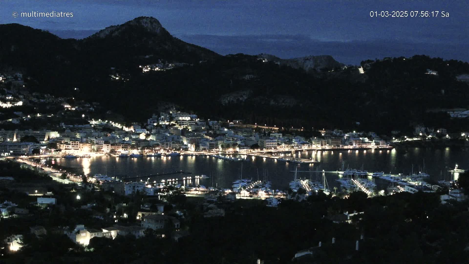 A nighttime view of a harbor town nestled between dark hills under a twilight sky.