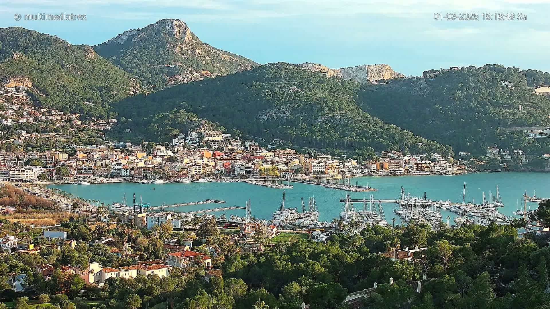 A coastal town nestled between green hills and a calm bay filled with sailboats, under a clear sky.