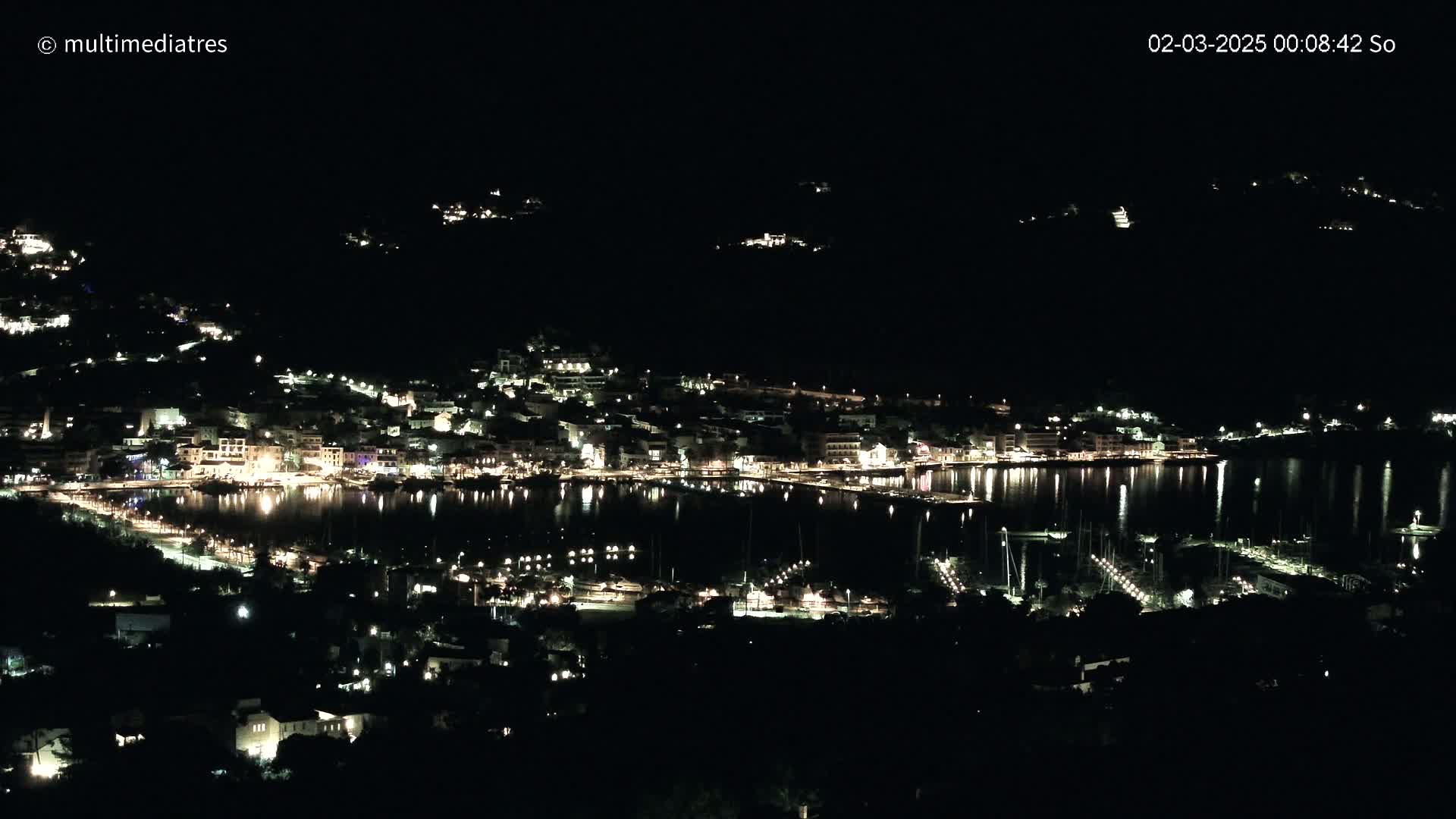 A nighttime, aerial view of a coastal town with its lights reflected on a calm body of water.