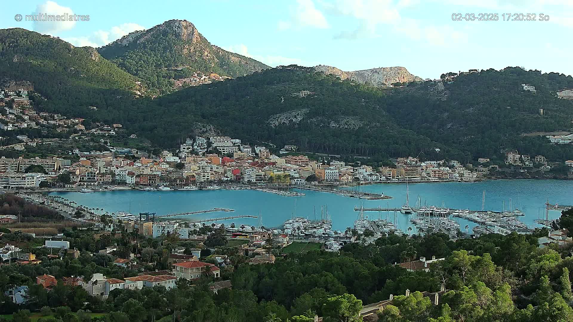 A coastal town nestled between mountains, with a marina filled with sailboats, under a partly cloudy sky.
