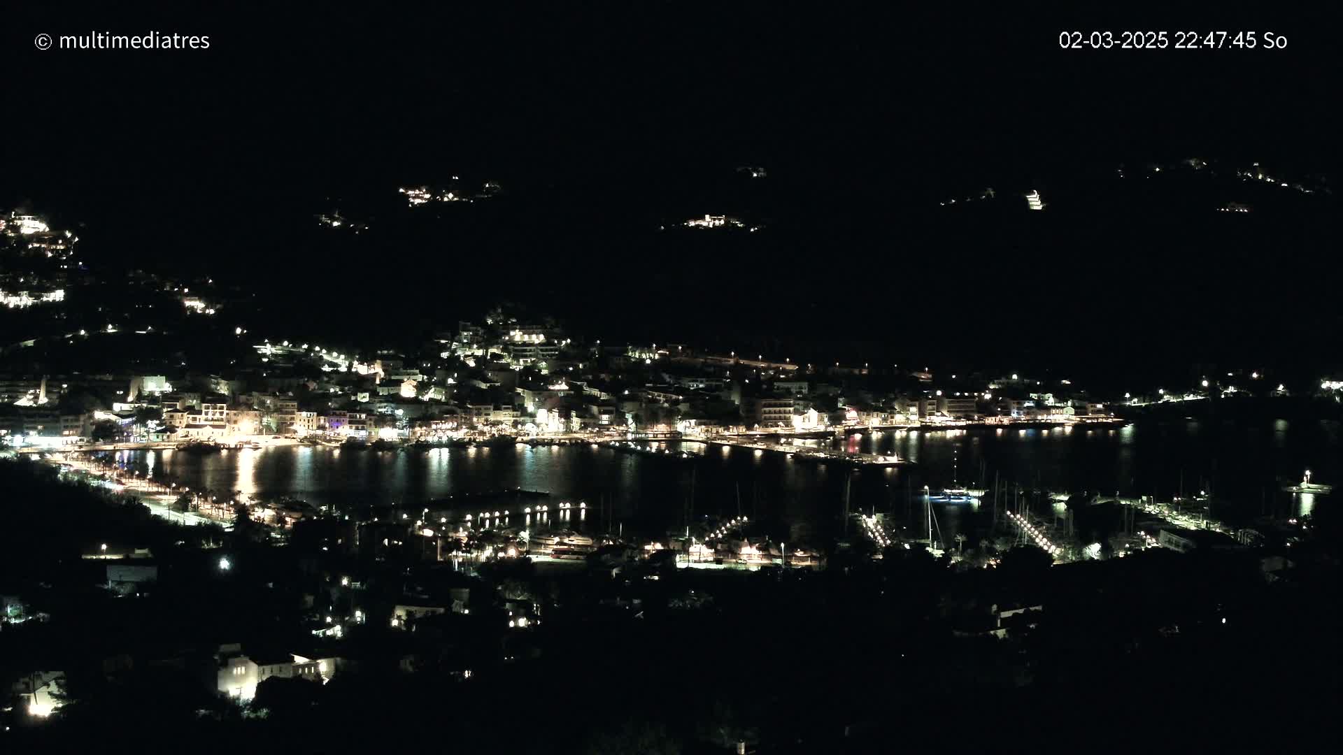 A nighttime view of a coastal town with many lights illuminating buildings and a harbor filled with boats.