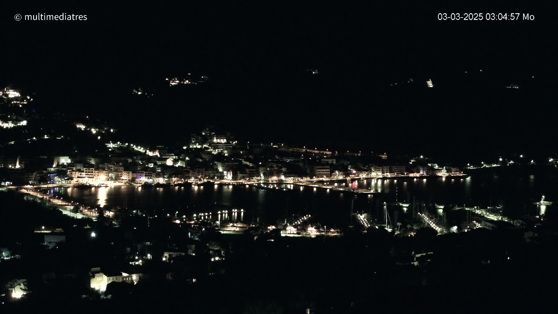 A nighttime, high-angle view shows a coastal town with many lights illuminating buildings and a harbor filled with boats.