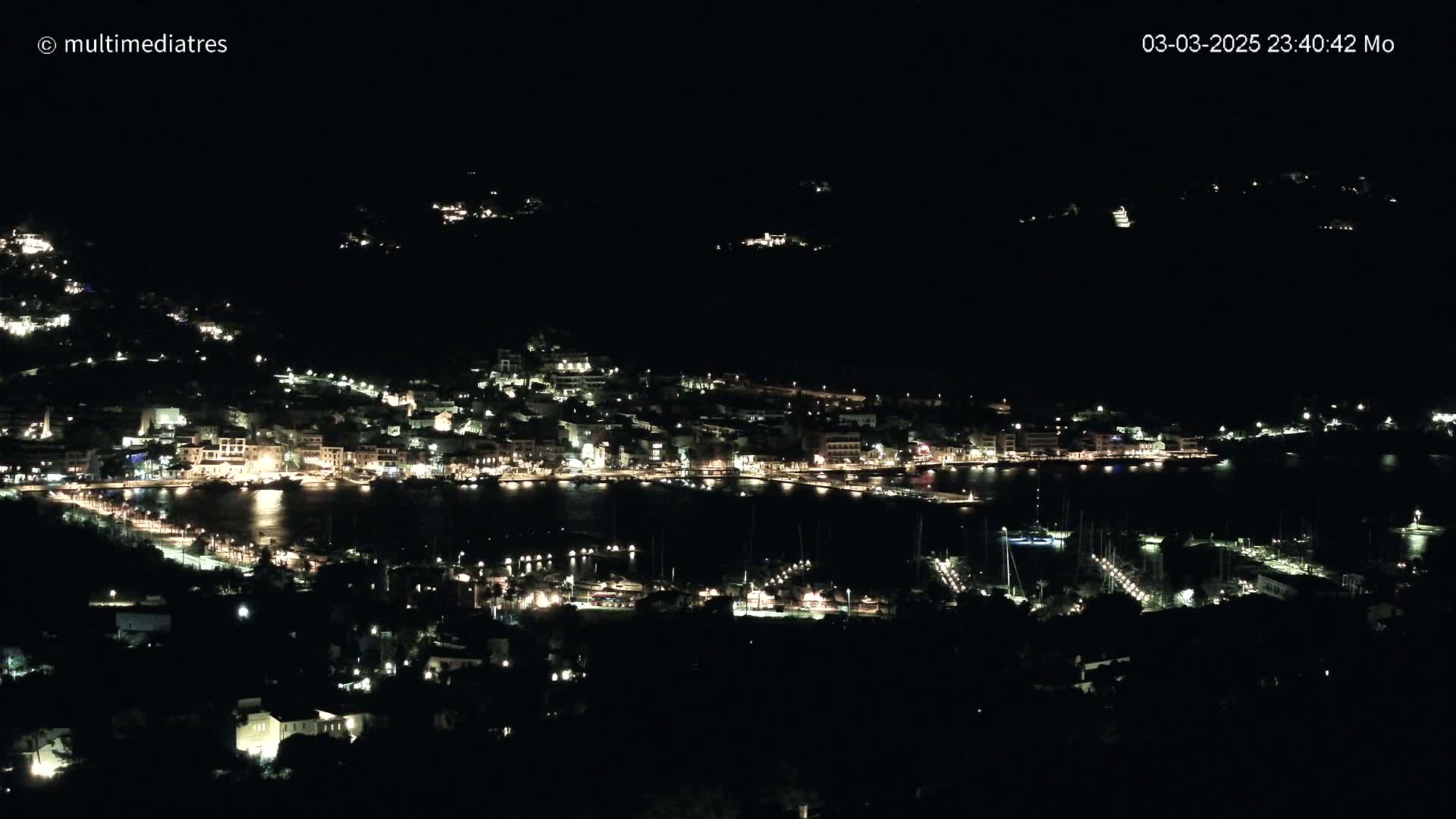 A coastal town at night, illuminated by many lights, is seen from a high vantage point under a dark, clear sky.
