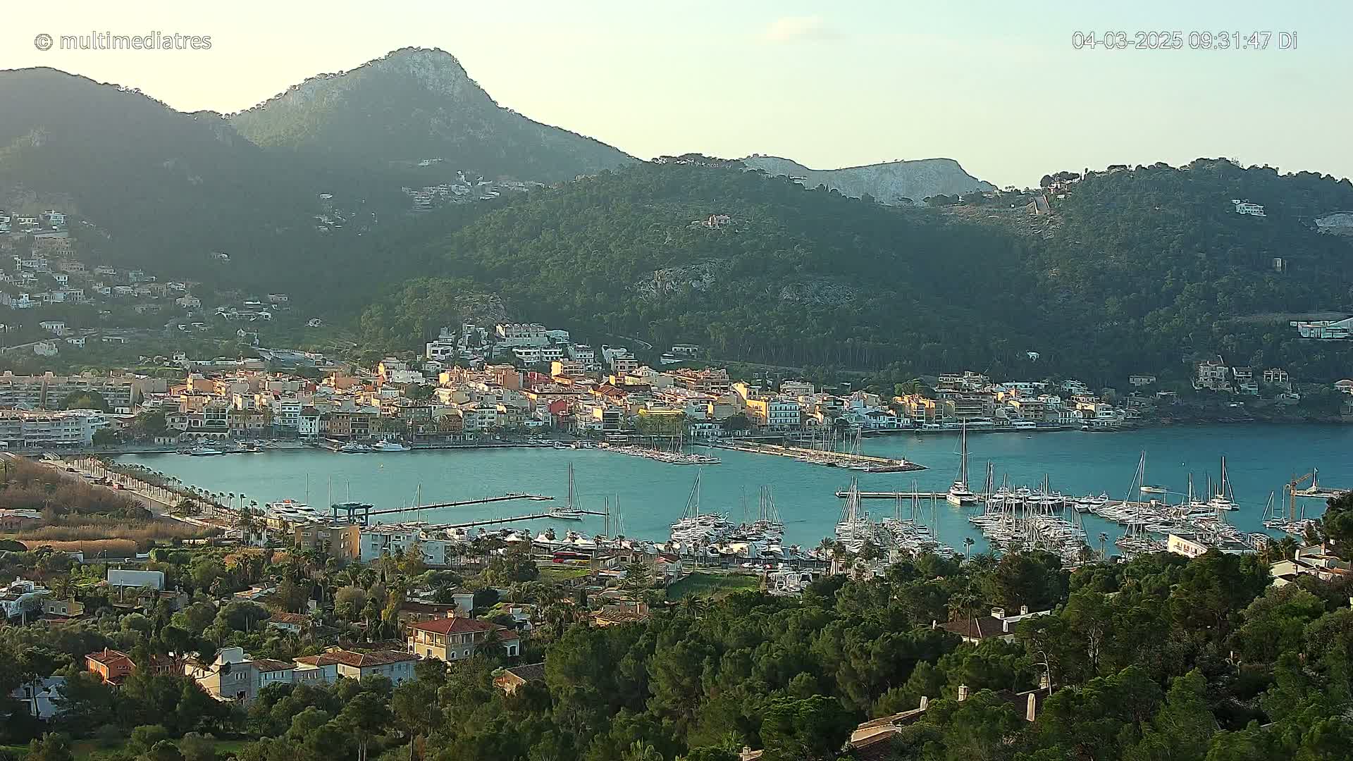 A coastal town nestled between green hills and a calm bay filled with numerous sailboats and yachts under a clear sky.
