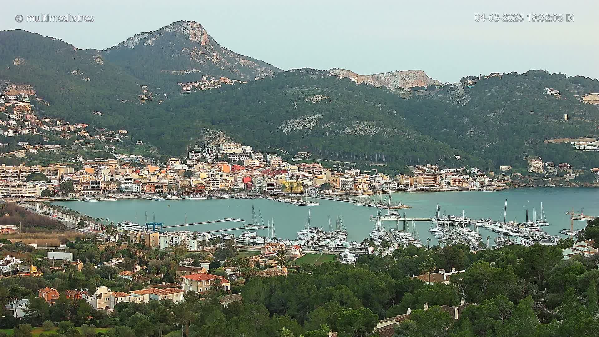 A coastal town with a marina filled with boats is nestled at the base of a mountainous region under an overcast sky.