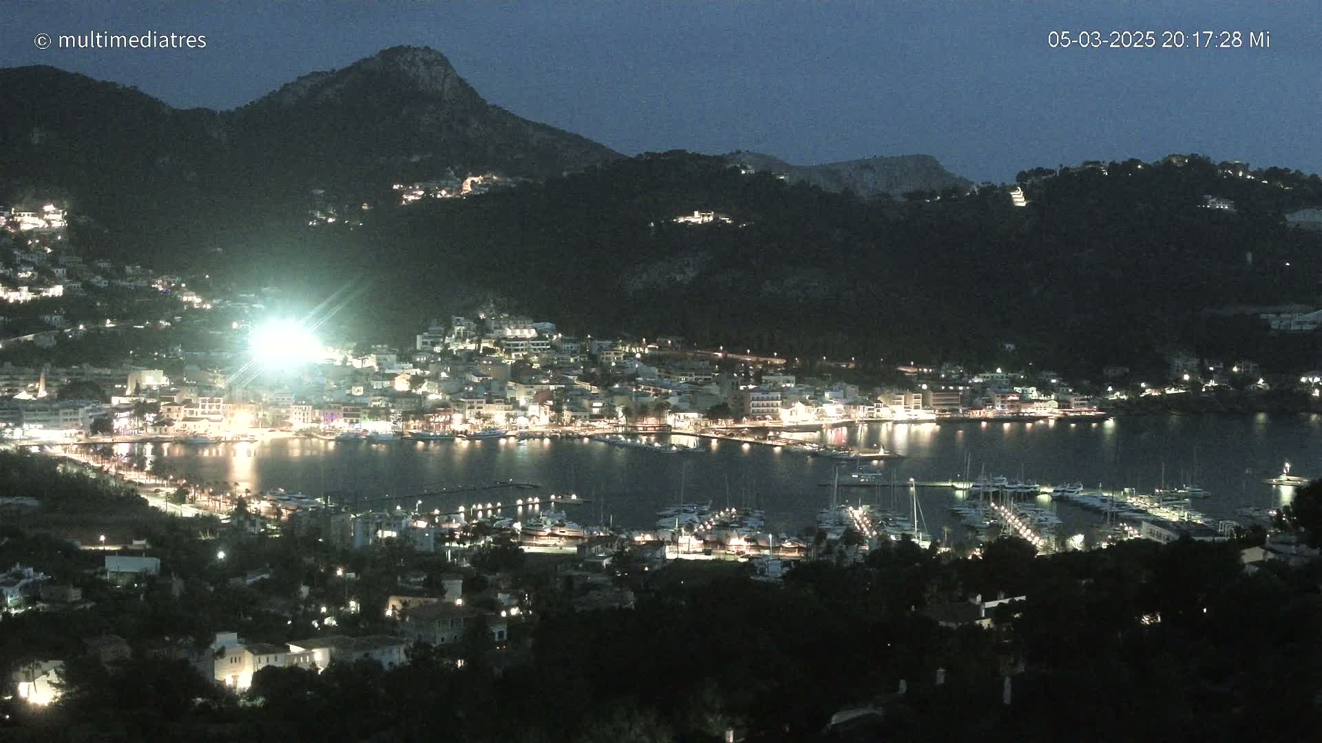 A nighttime view of a harbor town nestled between hills, illuminated by lights, under a dark, clear sky.
